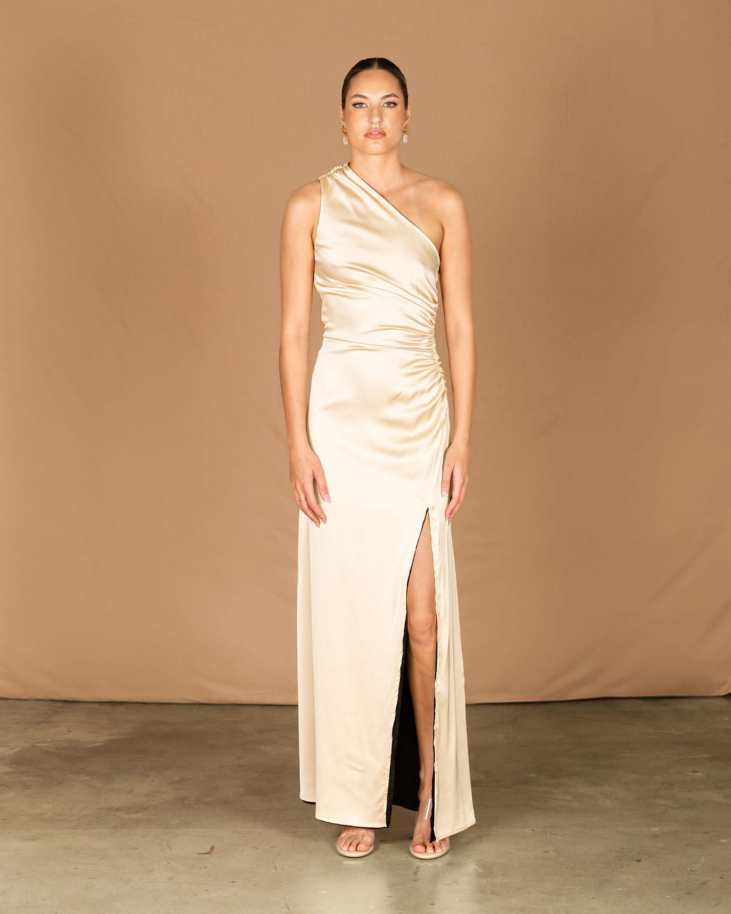 Woman wearing a one-shoulder beige evening gown with a slit on a brown background